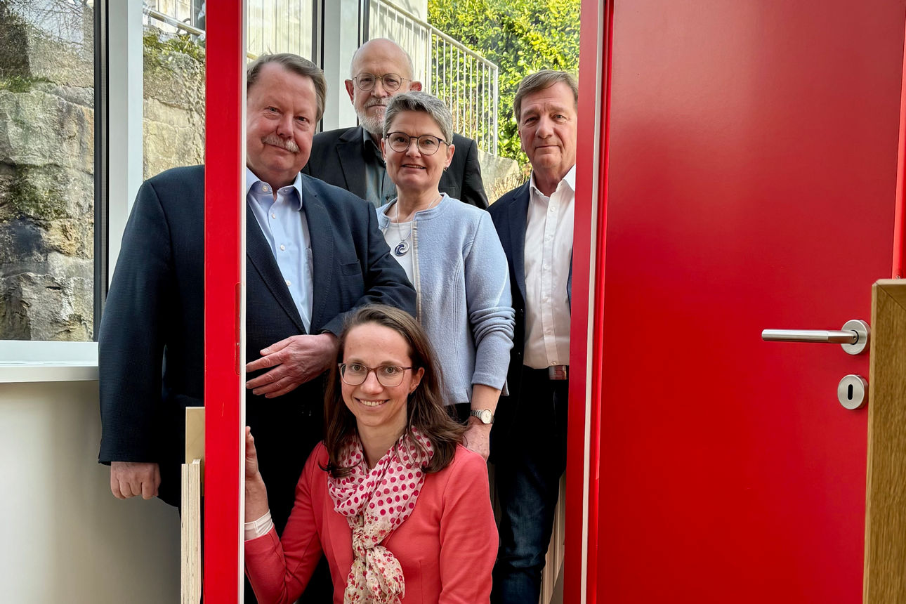 Das Team der Ketteler-Stiftung schaut durch eine offene rote Tür. Davor eine Matte mit dem Schriftzug "Caritas öffnet Türen.