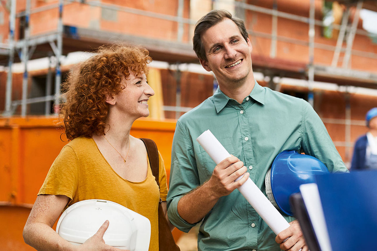 Mann und Frau stehen vor Baustelle mit Bauplan und Baufiberaterin