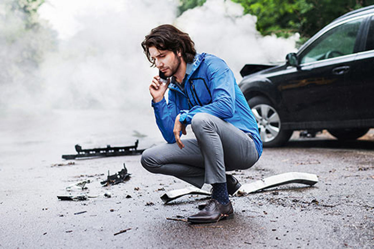 Ein Mann hockt vor einem beschädigten Auto und telefoniert