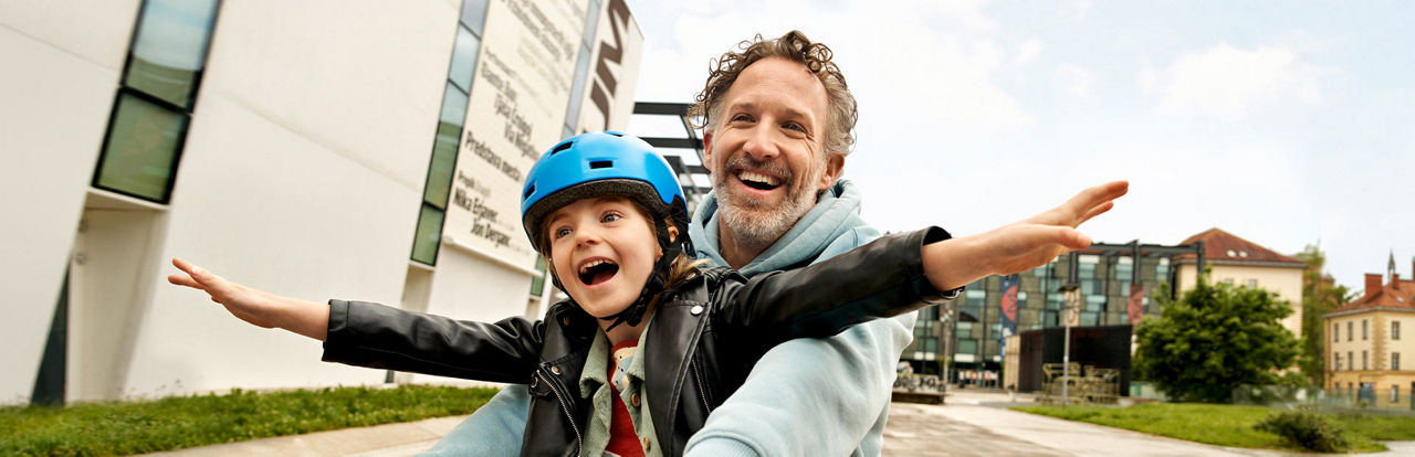 Kind und Vater fahren gemeinsam Fahrrad, während das Kind jubelnd die Hände hochhält