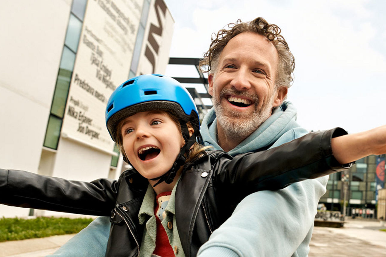 Kind und Vater fahren gemeinsam Fahrrad, während das Kind jubelnd die Hände hochhält