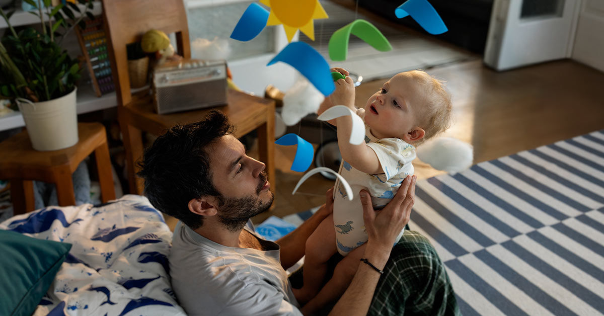 Ein junger Mann liegt auf dem Boden und spielt lächelnd mit einem Baby, das auf ihm liegt.