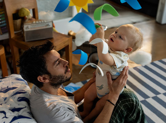 Ein junger Mann liegt auf dem Boden und spielt lächelnd mit einem Baby, das auf ihm liegt.