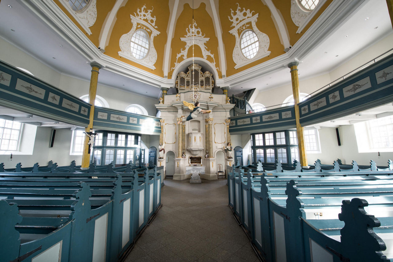 Blick in eine kleine Kirche mit Bänken. Am Altar schwebt eine Engelsfigur