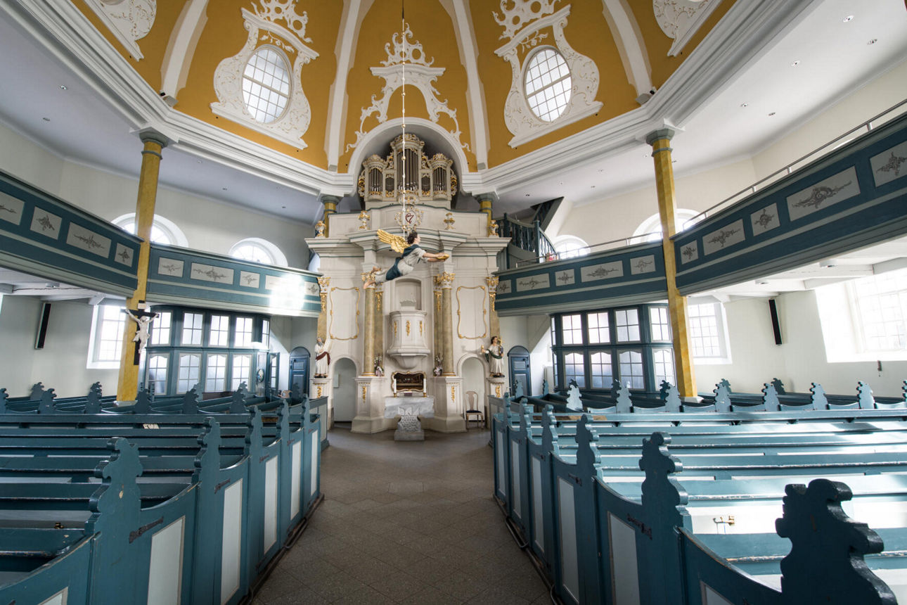 Blick in eine kleine Kirche mit Bänken. Am Altar schwebt eine Engelsfigur