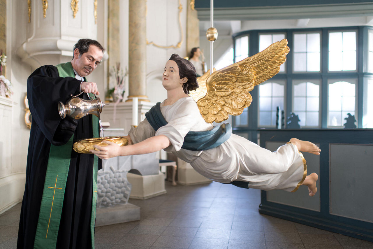 Ein Pastor steht in einer Kirche er gießt aus einem Krug Wasser in ein Taufbecken, das von einer Engelsfigur gehalten wird.
