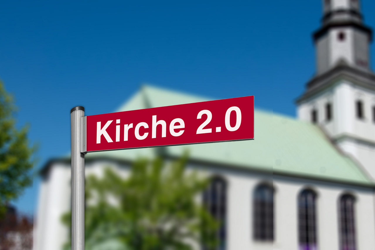 Schild mit der Aufschrift "Kirche 2.0" vor einer verschommenen Kirche