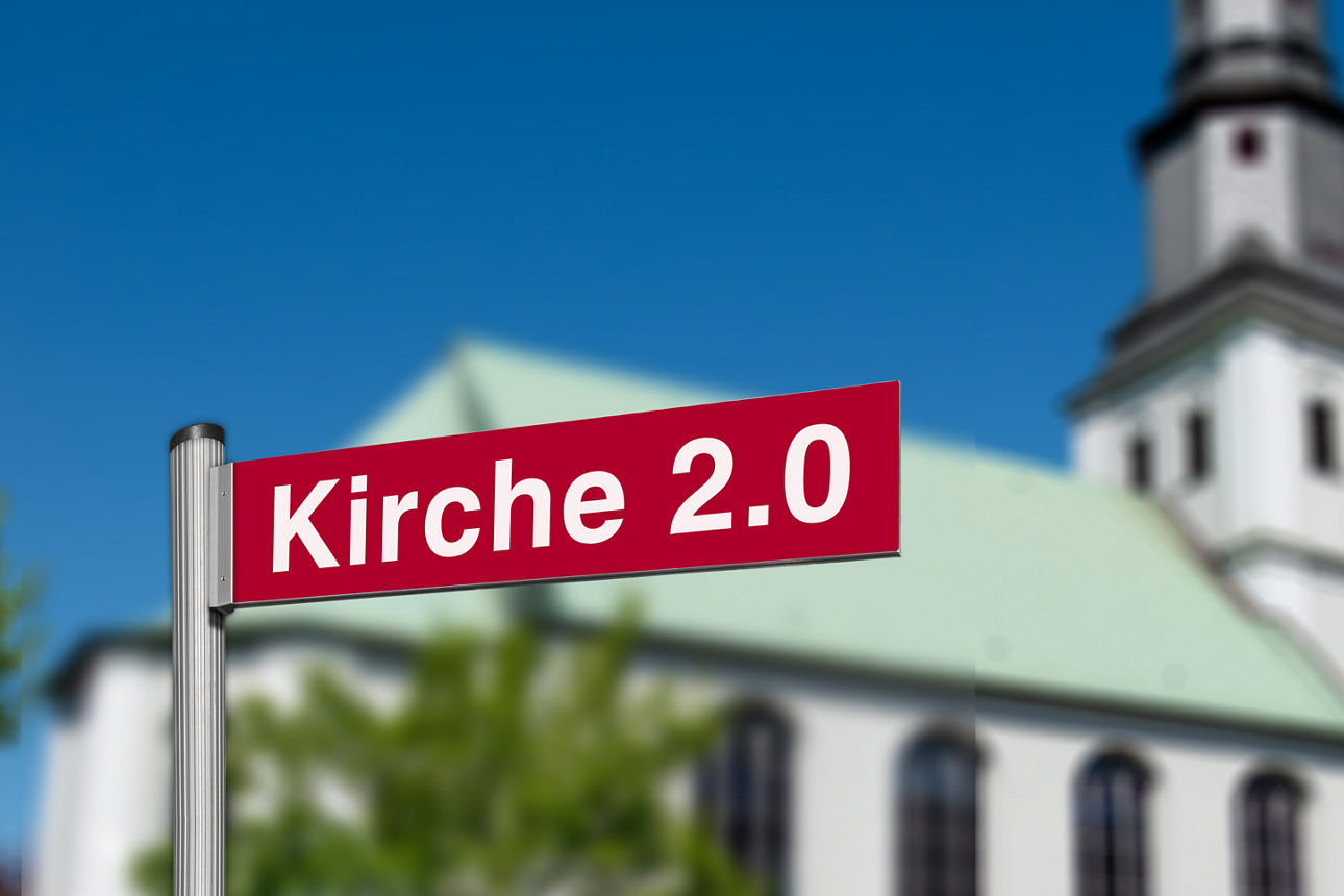 Schild mit der Aufschrift "Kirche 2.0" vor einer verschommenen Kirche
