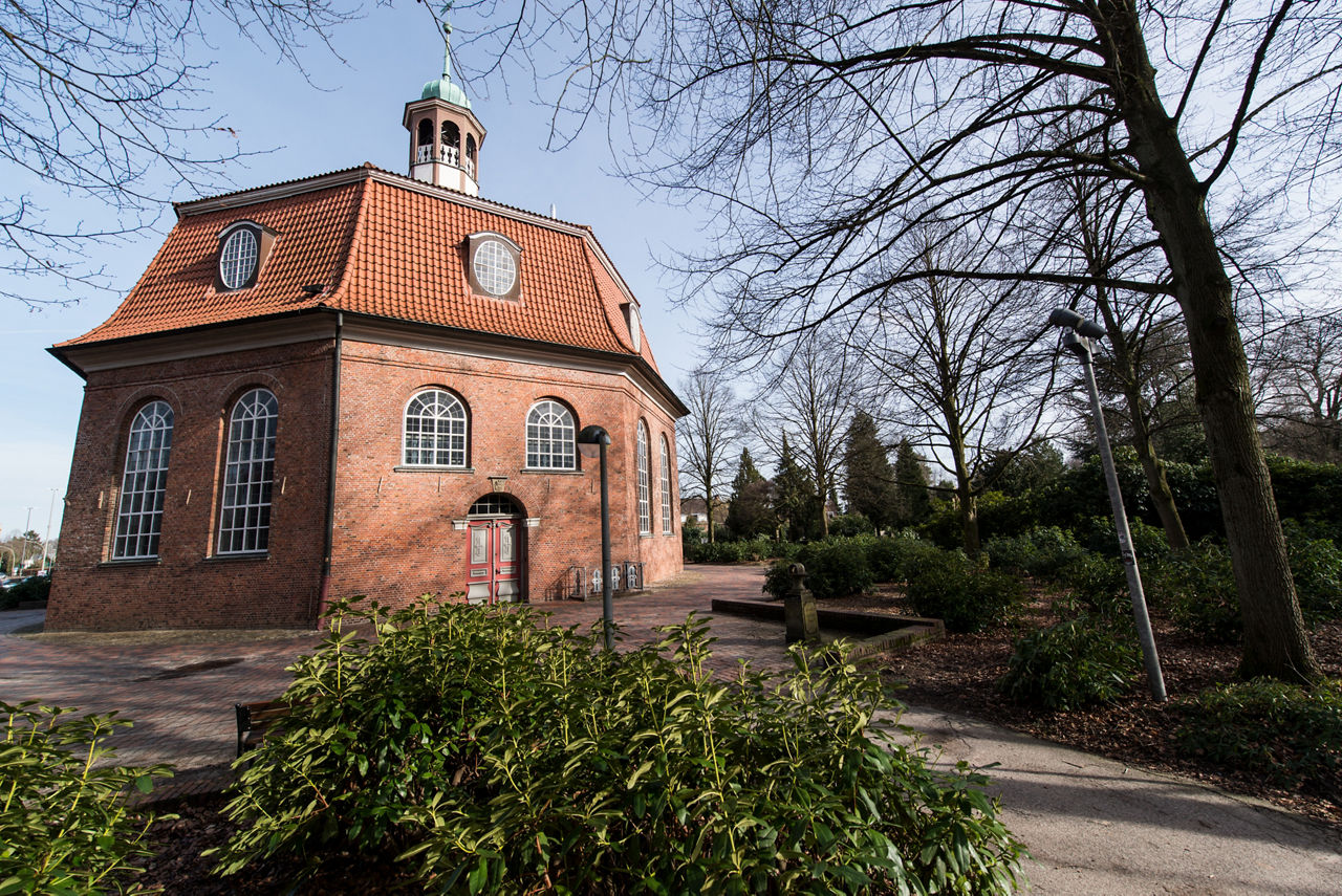 Backsteinkirche mit rotem Walmdach und mehreren hohen Rundbogenfenstern, umgeben von einem Park mit Büschen, Bäumen und gepfl