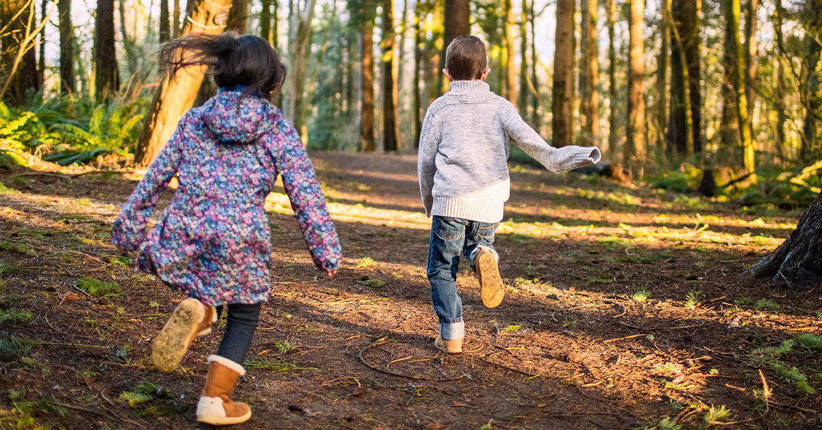 Zwei Kinder, ein Junge und ein Mädchen, rennen auf einem Waldweg. Es ist sonnig und sie tragen Herbstkleidung.