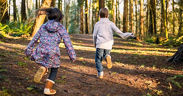 Zwei Kinder, ein Junge und ein Mädchen, rennen auf einem Waldweg. Es ist sonnig und sie tragen Herbstkleidung.