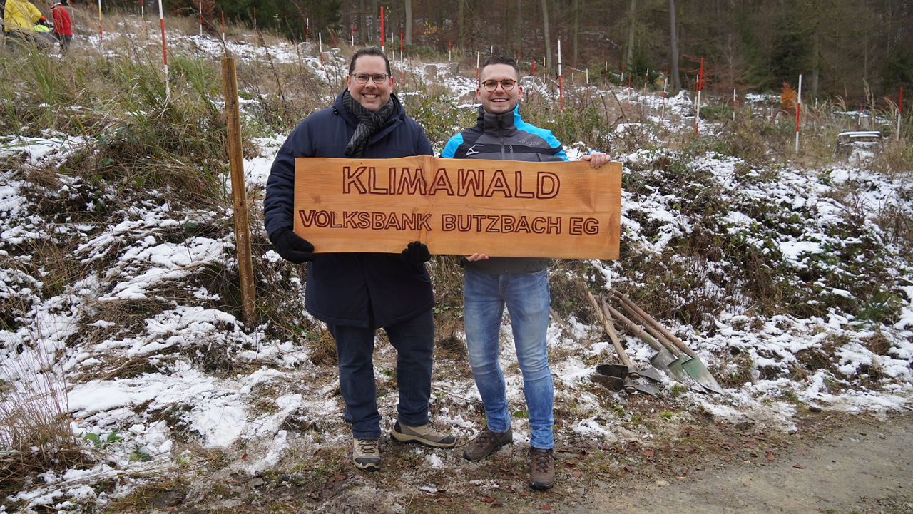 Zwei Personen halten ein Holzschild auf dem Klimawald Volksbank Butzbach eG steht in einem Wald