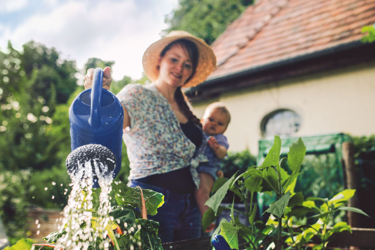Eine Frau mit Baby auf dem Arm gießt mit einer blauen Gießkanne ein Hochbeet