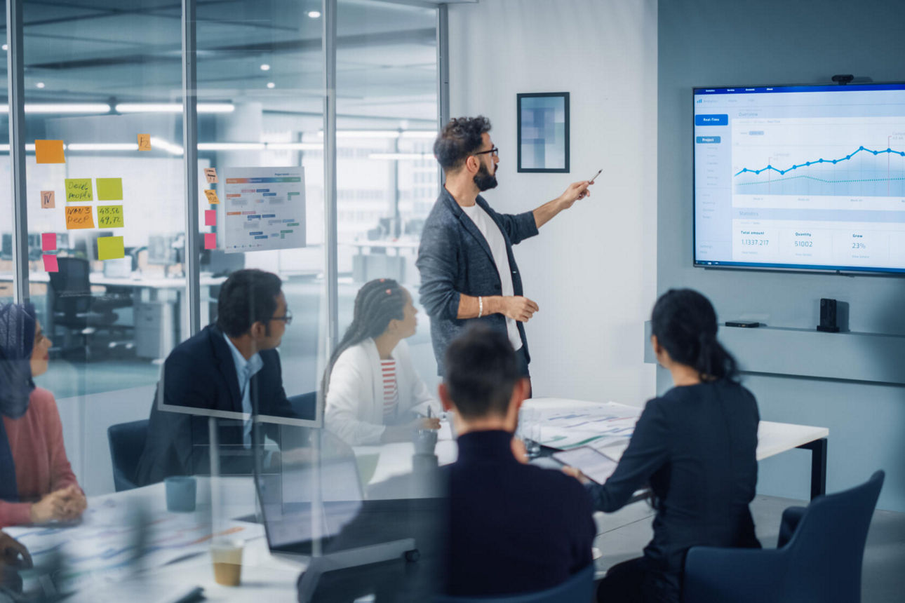 In einem Konferenzraum hält ein Mann eine Präsentation und erklärt einen Graphen auf dem Monitor.