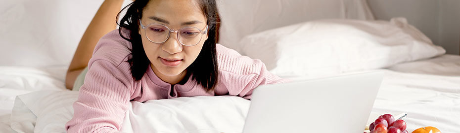 Eine junge Frau mit langen schwarzen Haaren liegt auf einem Bett, trägt einen rosa Pullover und eine Brille.