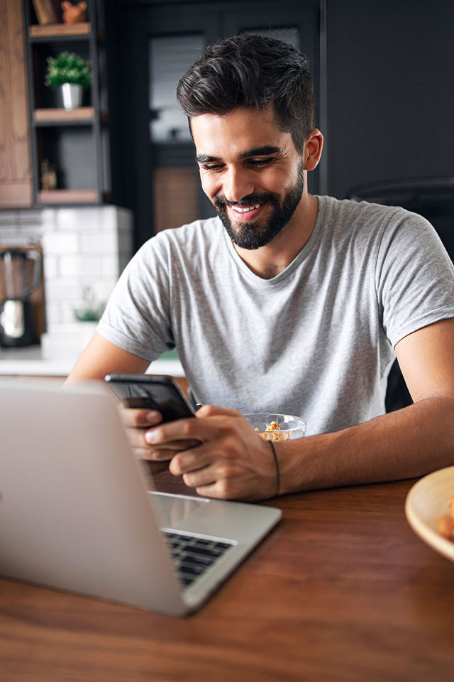 Ein junger Mann sitzt an einem Tisch mit einem Laptop und schaut lächelnd auf ein Smartphone