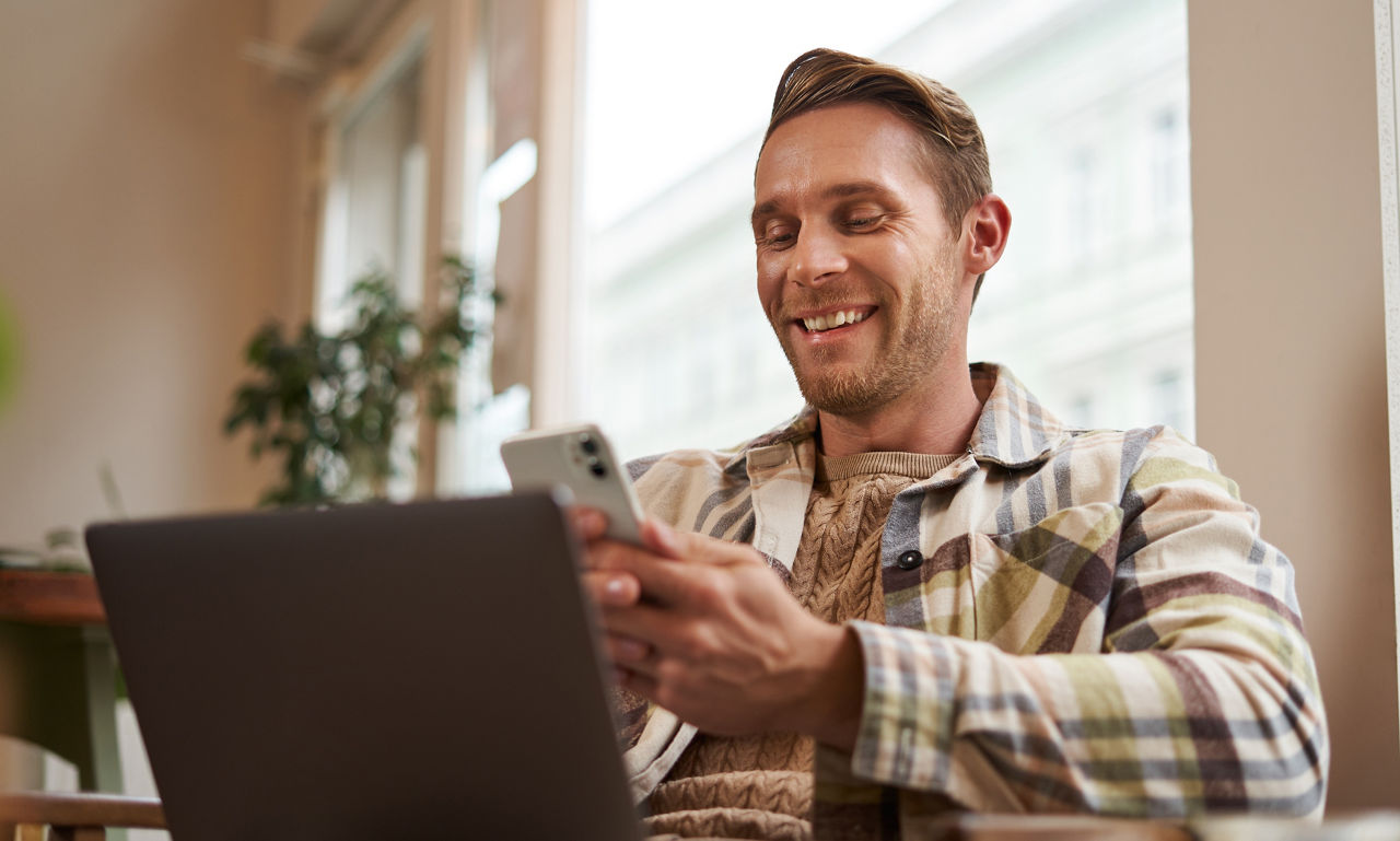 Portrait eines glücklichen Mannes mit Handy sitzend vor Laptop