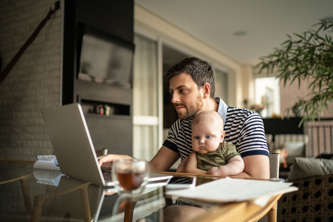 Ein Mann sitzt am Küchentisch und hat dabei sein Baby auf dem Schoß, welches er mit seinem linken Arm festhält. Vor ihm auf dem Tisch steht ein geöffneter Laptop, den er mit seiner rechten Hand bedient.