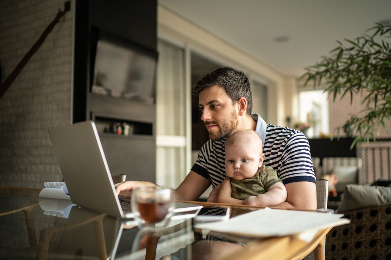Ein Mann sitzt am Küchentisch und hat dabei sein Baby auf dem Schoß, welches er mit seinem linken Arm festhält. Vor ihm auf dem Tisch steht ein geöffneter Laptop, den er mit seiner rechten Hand bedient.