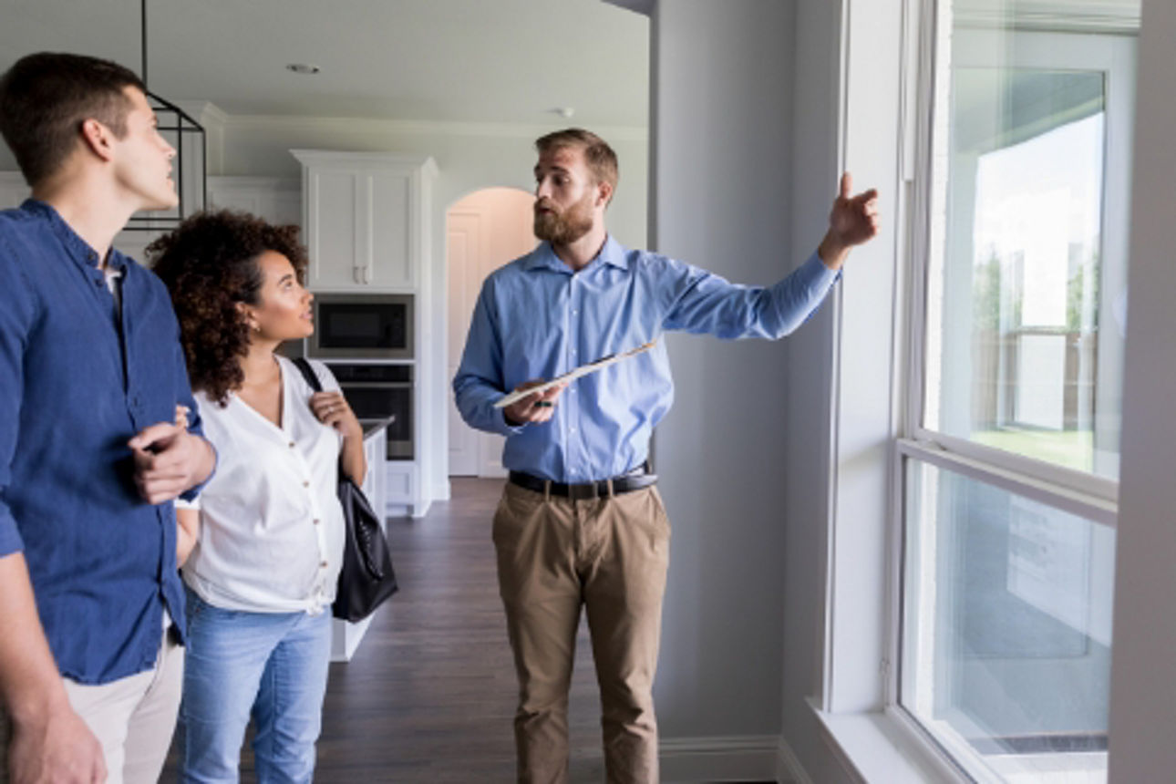Ein Paar wird in einer leerstehenden Wohnung beraten, der Berater zeigt auf die Fenster