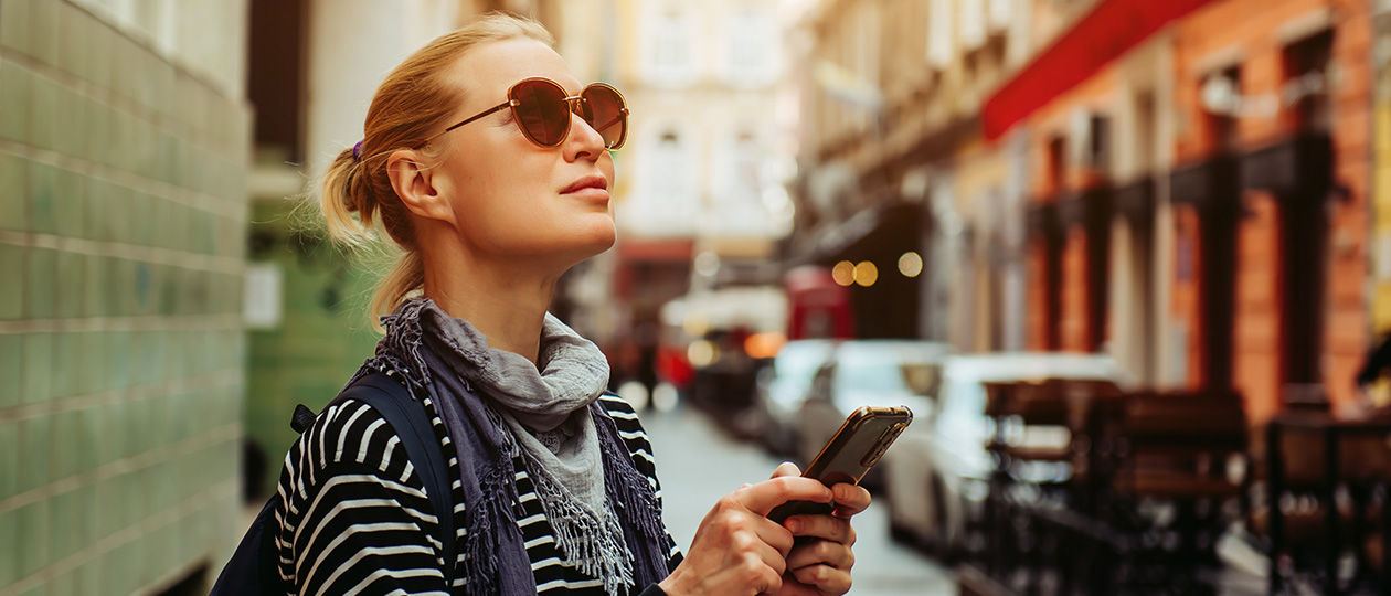 junge Frau mit Sonnenbrille steht in der Stadt und schaut aufs Handy