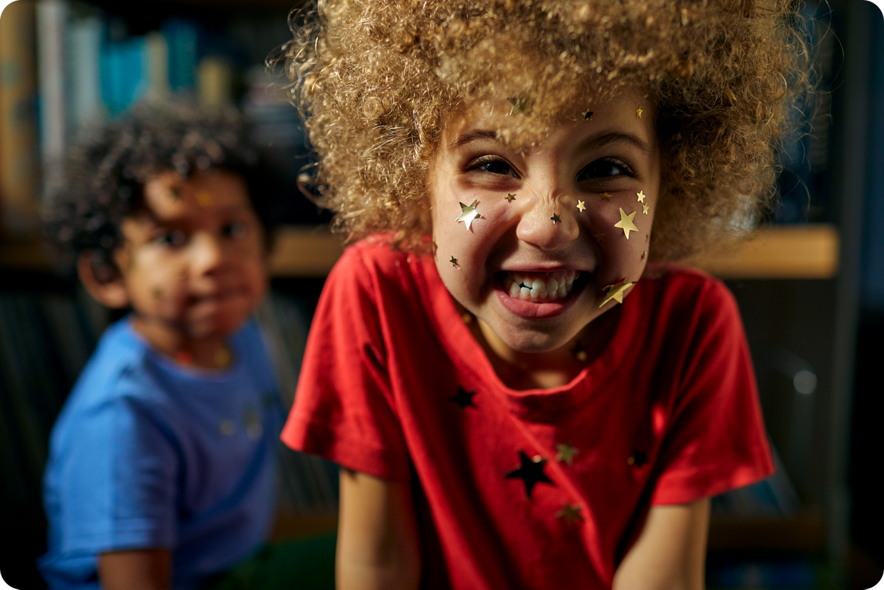 Kleines Mädchen mit einem Lockenkopf grinst in die Kamera und hat das Gesicht voller Sticker