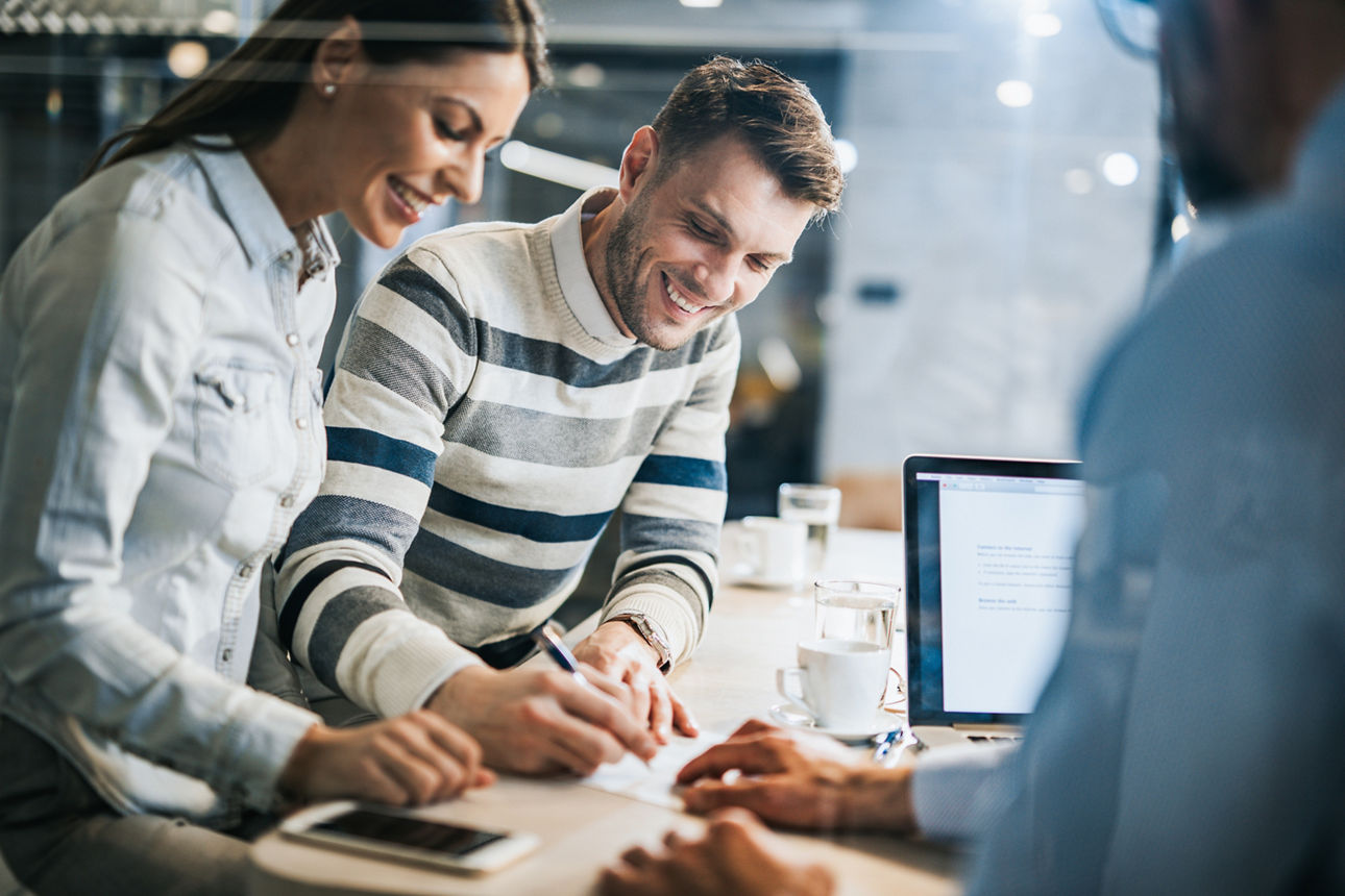 Kredit aufnehmen Ein junges, glückliches Paar unterzeichnet ein Dokument während eines Meetings mit ihrer Immobilienmaklerin im Büro. Der Blick erfolgt durch eine Glasscheibe.