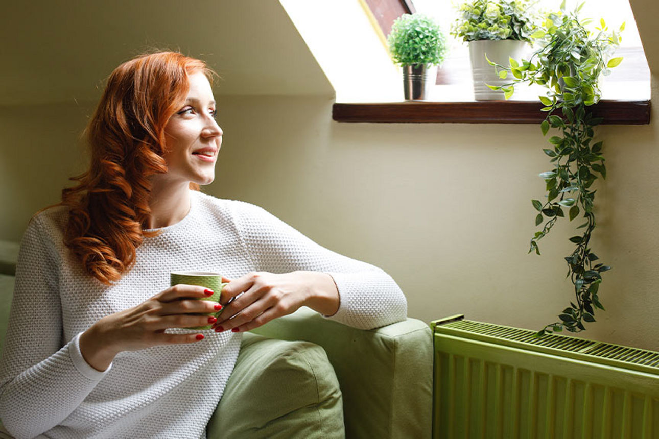 Junge Frau sitzt lächelnd mit Kaffeetasse vor ihrer Heizung