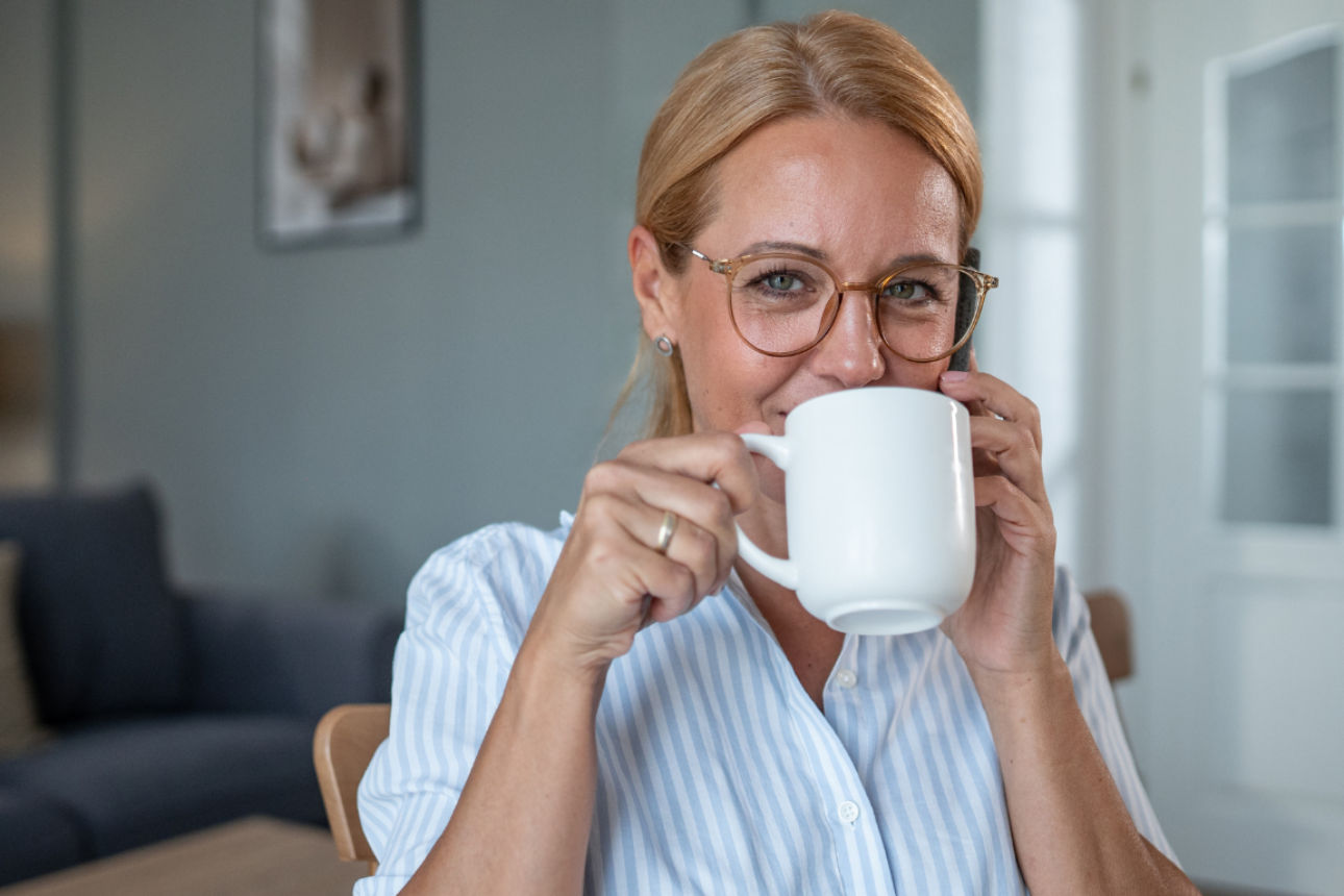 Person sitzt zu Hause und trinkt Kaffee aus einer weißen Tasse, im Hintergrund Wohnzimmer mit Sofa und Regal.