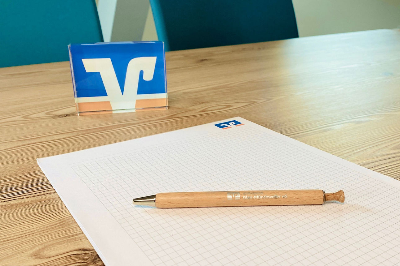 Ein gebrandeter Kugelschreiber in Holzoptik liegt auf einem Volksbank Papierblock. Beide liegen auf einem Holtisch.