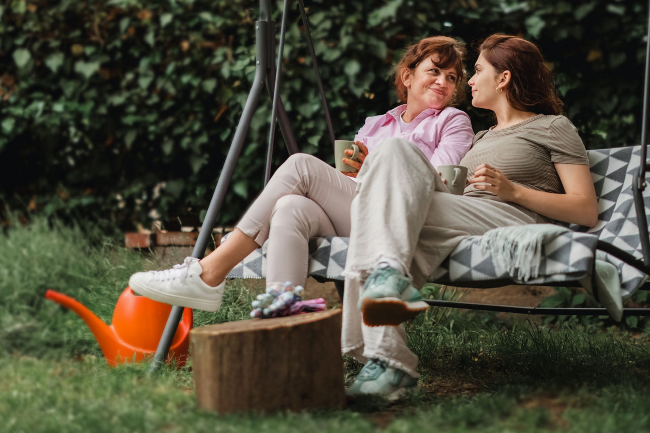 Zwei Frauen sitzen entspannt mit Tassen auf einer Gartenschaukel und unterhalten sich im Grünen.