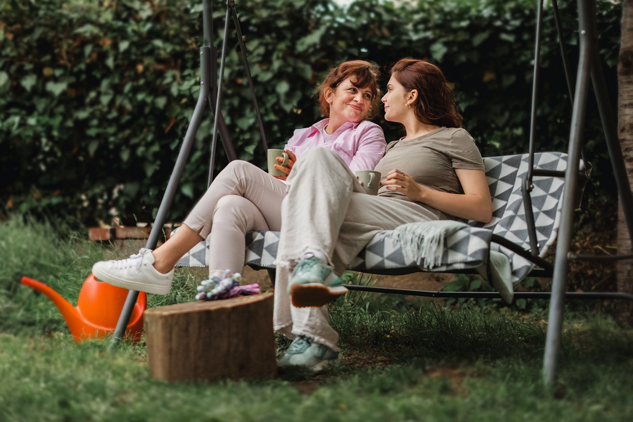 Zwei Frauen sitzen entspannt mit Tassen auf einer Gartenschaukel und unterhalten sich im Grünen.