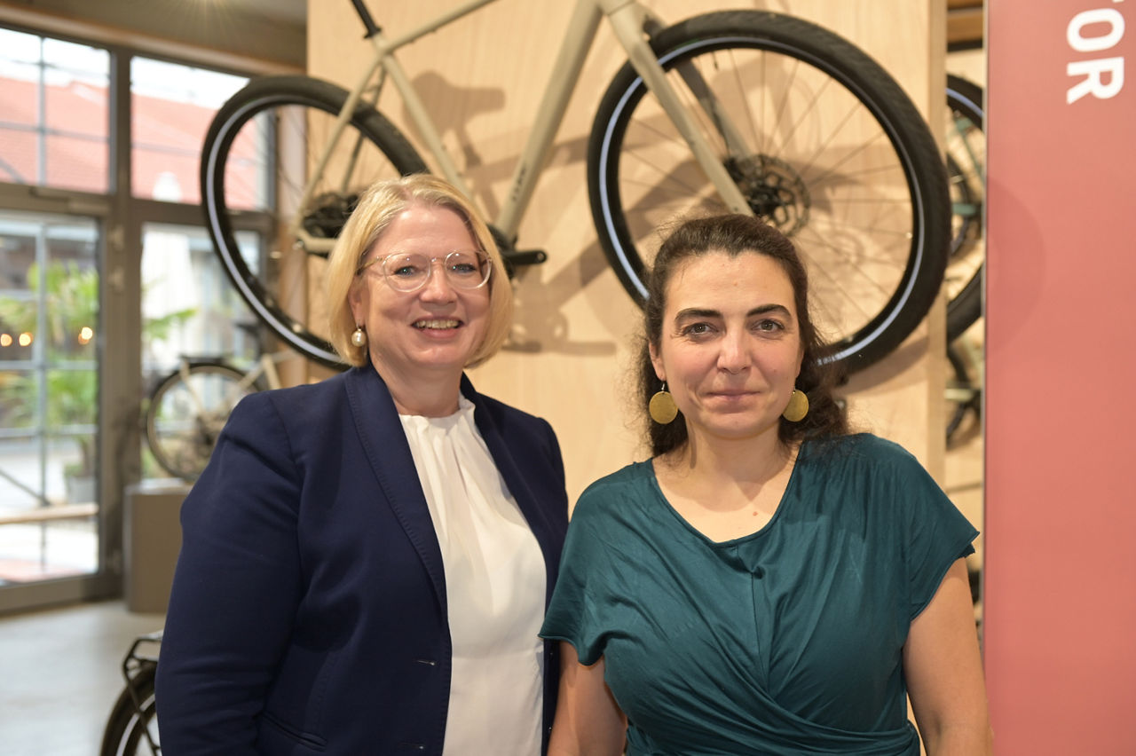 Zwei Damen stehen in einem Fahrradgeschäft vor einer Wand mit einem aufgehängtem Mountainbike, im Hintergrund der Laden mit großen Fenstern ins Freie