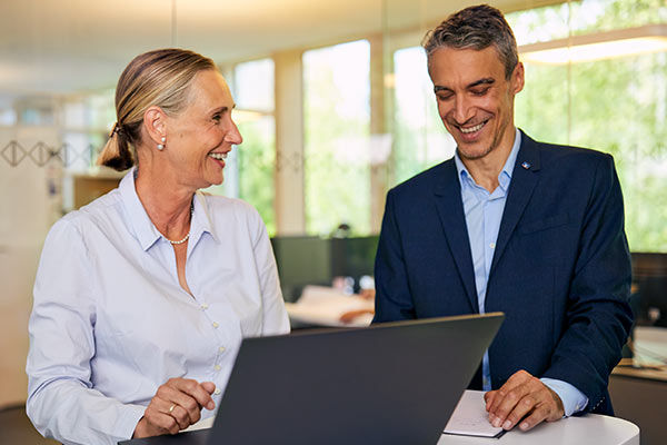 Eine Frau und ein Mann in Businesskleidung unterhalten sich lächelnd vor einem Laptop in einem modernen Büro.