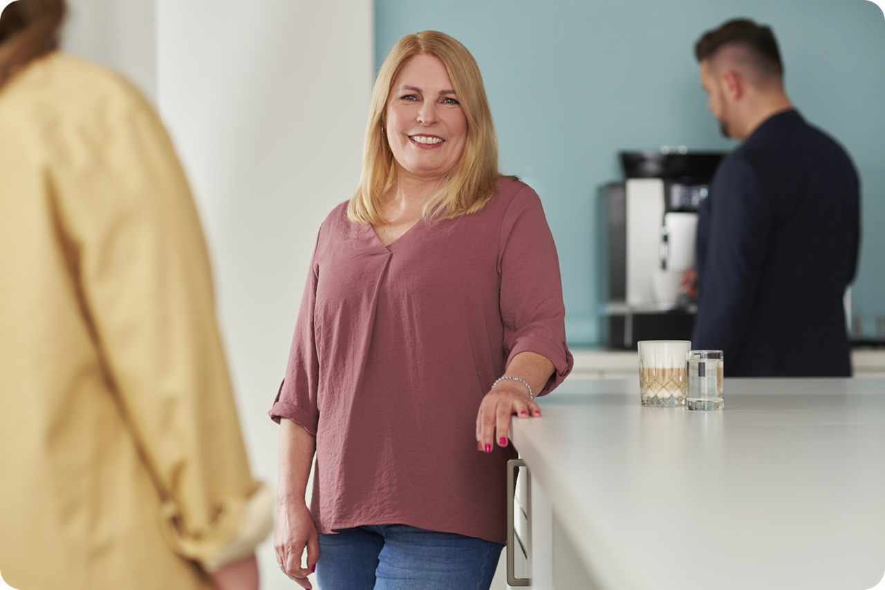 Eine Frau steht an einem Sideboard und lächelt in die Kamera, neben ihr steht eine Tasse Kaffee