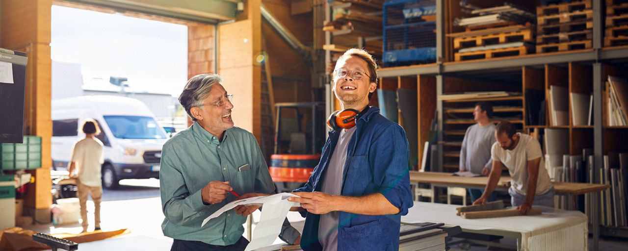 Älterer und jüngerer Mann stehen in einer Arbeitshalle und besprechen Arbeitsdokumente