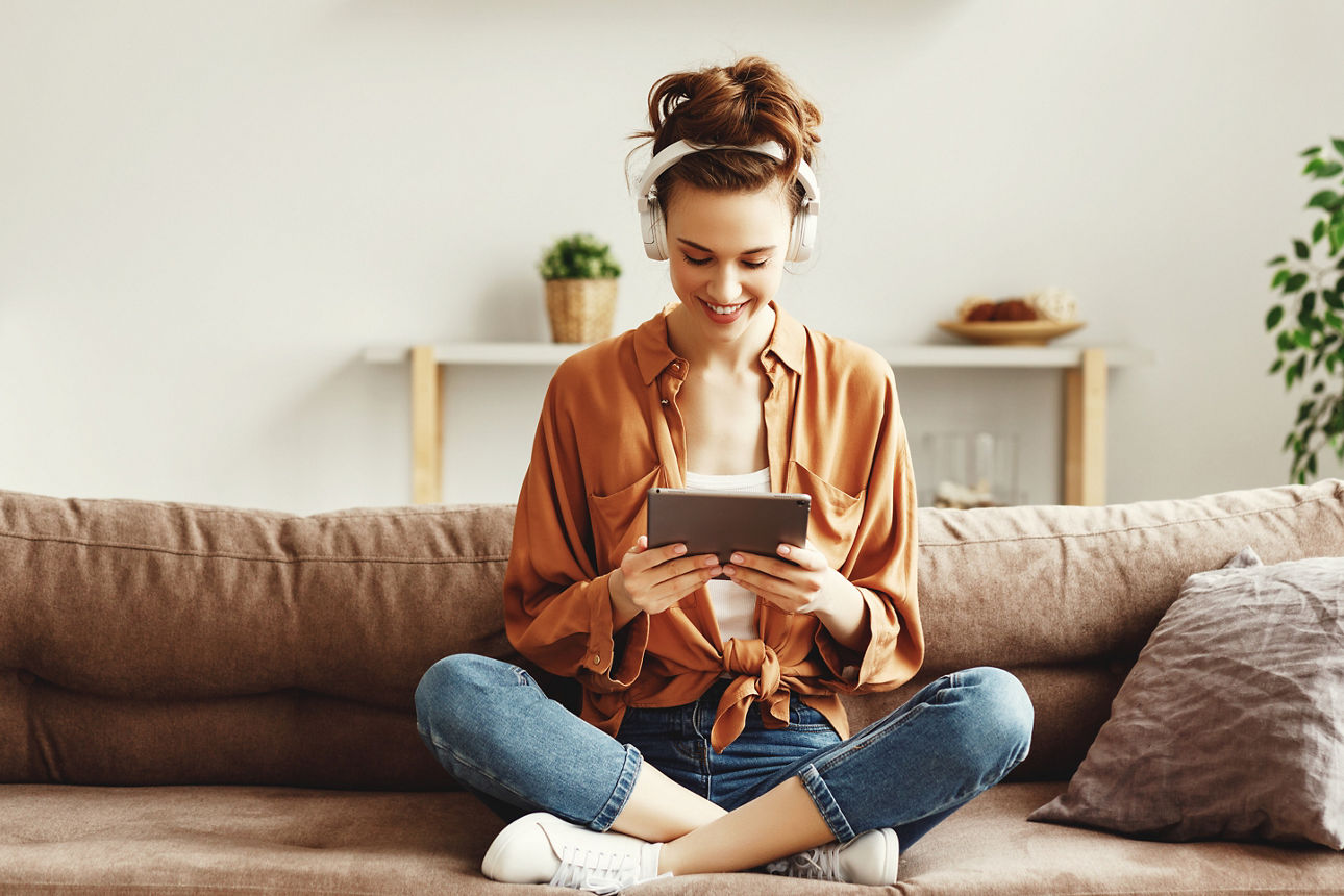 Eine Frau sitzt mit Kopfhörer und Tablet im Schneidersitz auf einer Couch