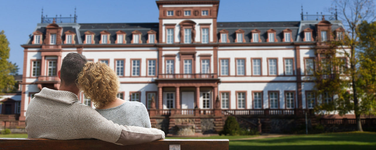Paar sitzt eng umschlungen auf einer Parkbank vor einem Schloss