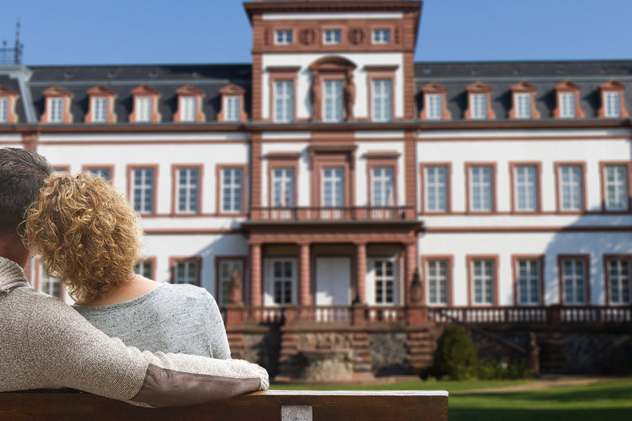 Paar sitzt eng umschlungen auf einer Parkbank vor einem Schloss