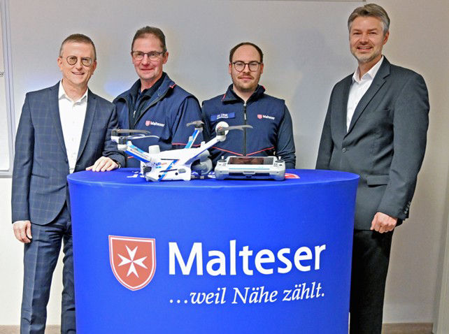 Vier Männer stehen hinter einem blauen Stehtisch mit der Aufschrift „Malteser ...weil Nähe zählt“, auf dem eine Drohne platzi
