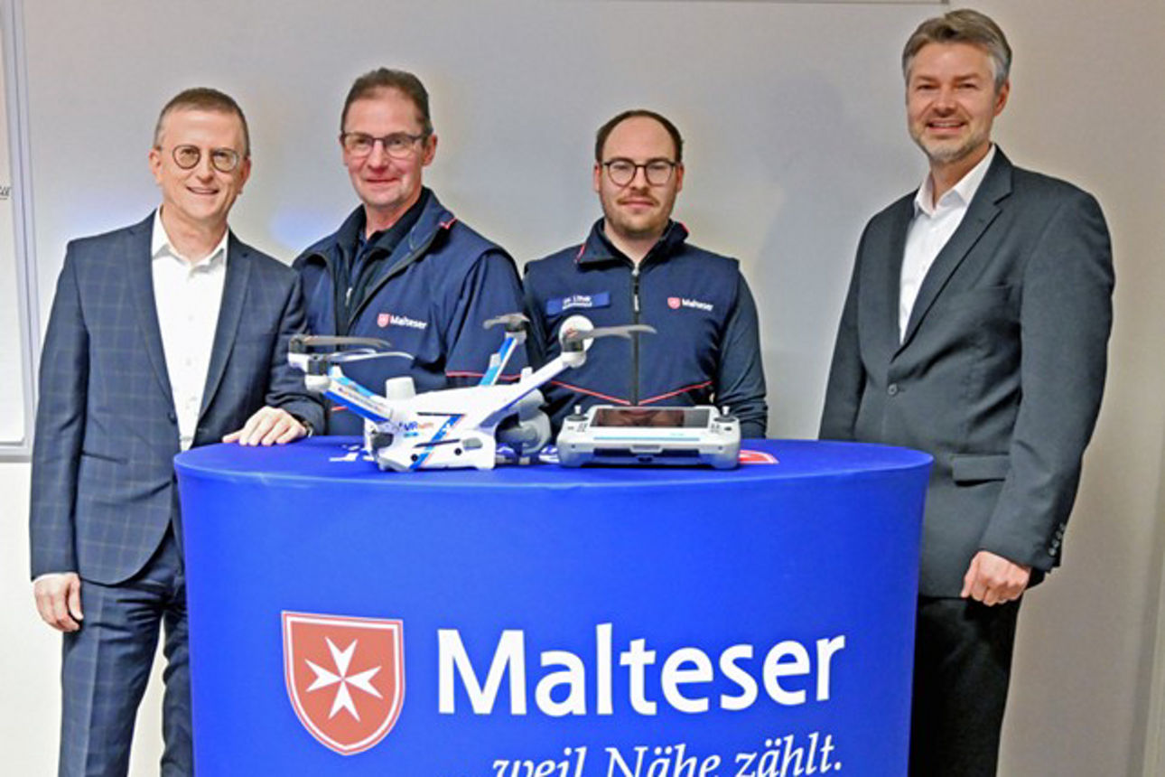 Vier Männer stehen hinter einem blauen Stehtisch mit der Aufschrift „Malteser ...weil Nähe zählt“, auf dem eine Drohne platzi