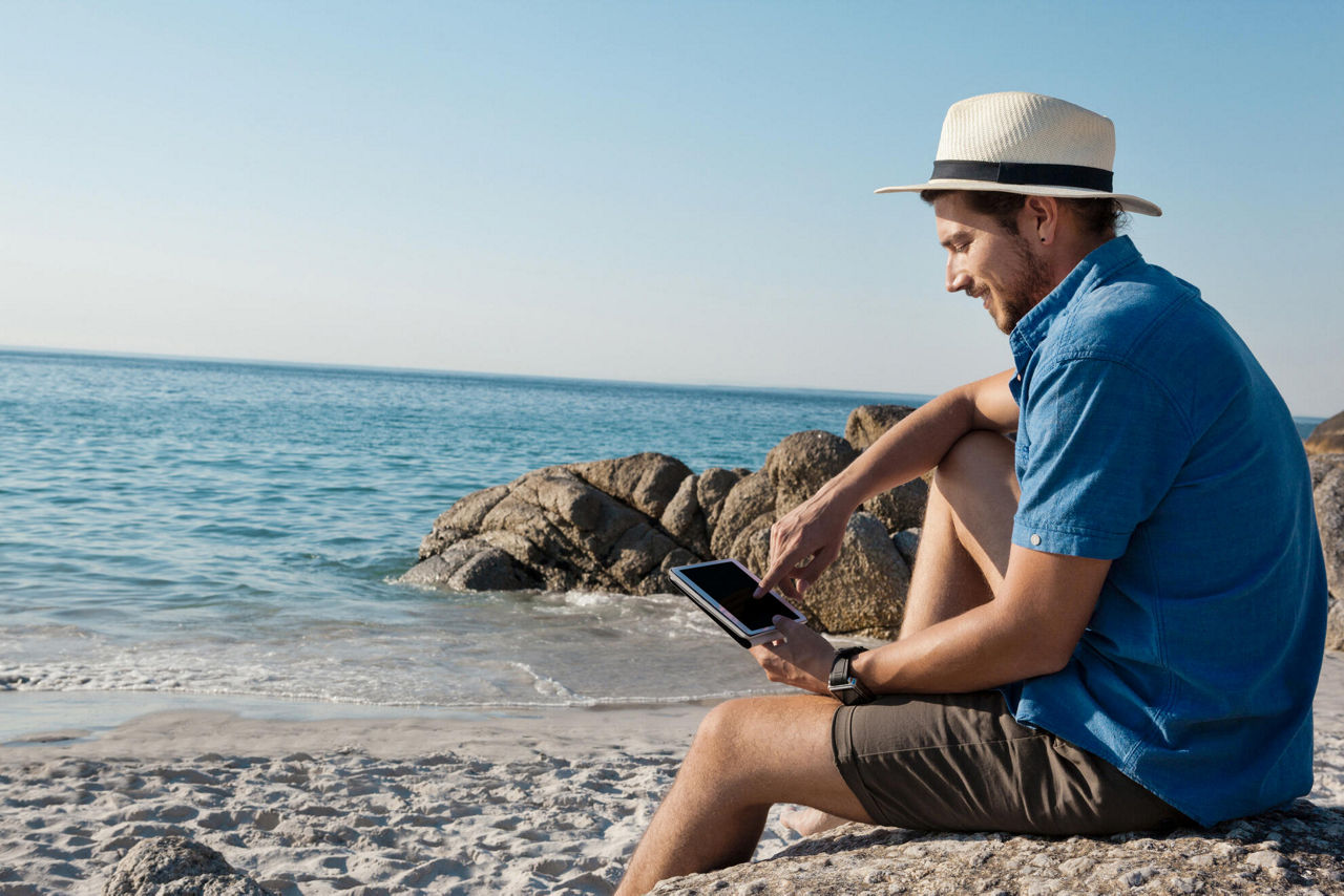 Ein junger Mann mit Hut sitzt am Strand und liest etwas auf seinem Tablet.