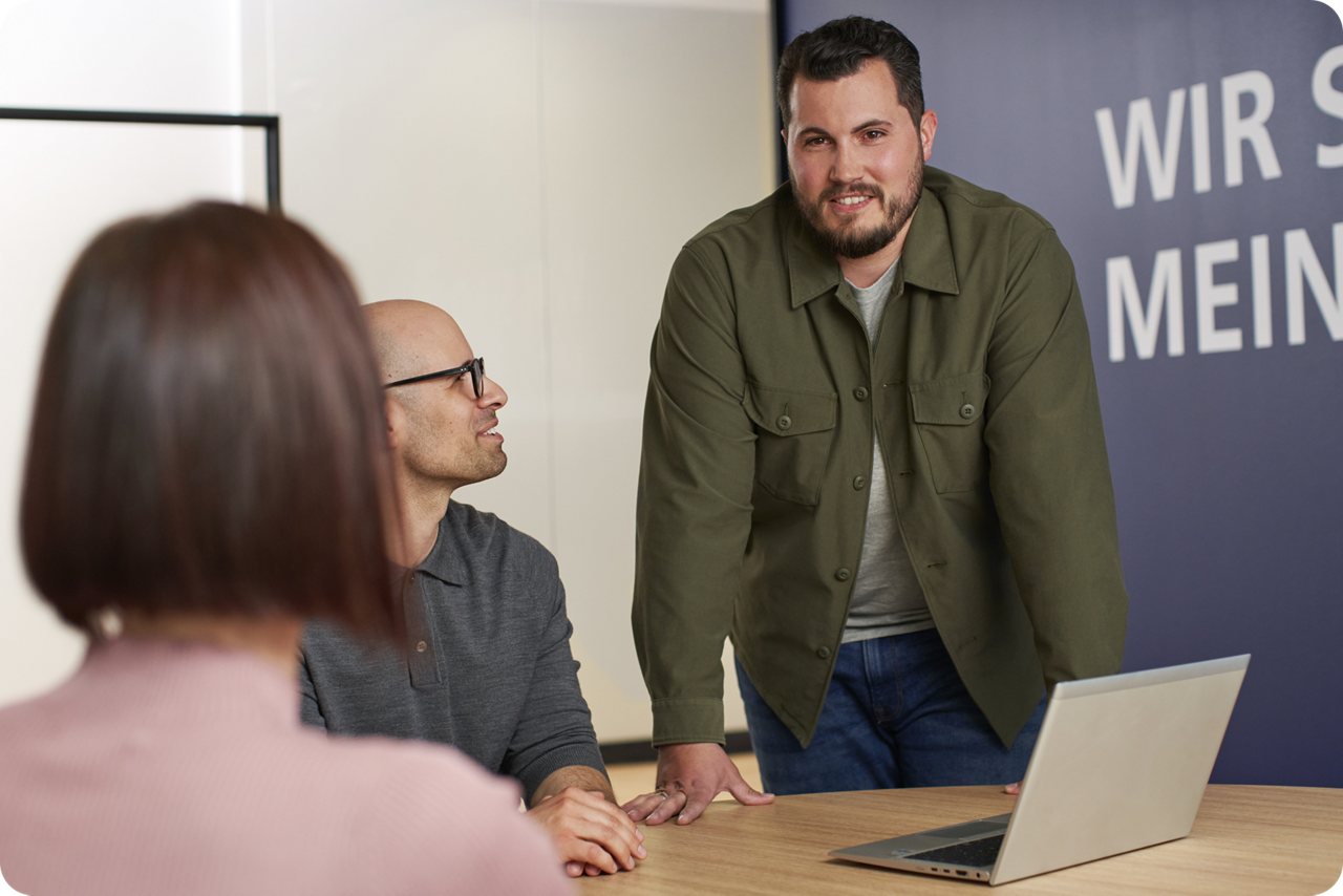 Zwei Personen sitzen an einem Tisch, eine Person steht, sie diskutieren miteinander