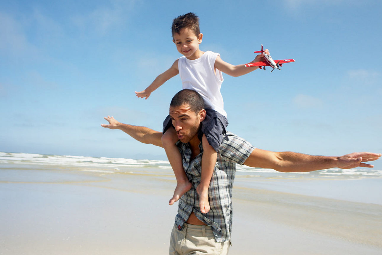 Ein Mann am Strand mit wie zum Fliegen ausgebreiteten Armen trägt ein Kind mit einem Spielzeugflieger auf den Schultern
