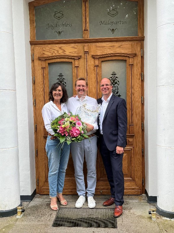 Eine Frau und zwei Männer stehen strahlend vor einer Eingangstür. Die Frau hält einen bunten Blumenstrauß in den Händen.