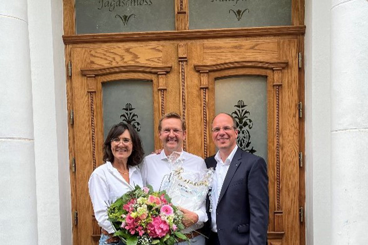 Eine Frau und zwei Männer stehen strahlend vor einer Eingangstür. Die Frau hält einen bunten Blumenstrauß in den Händen.