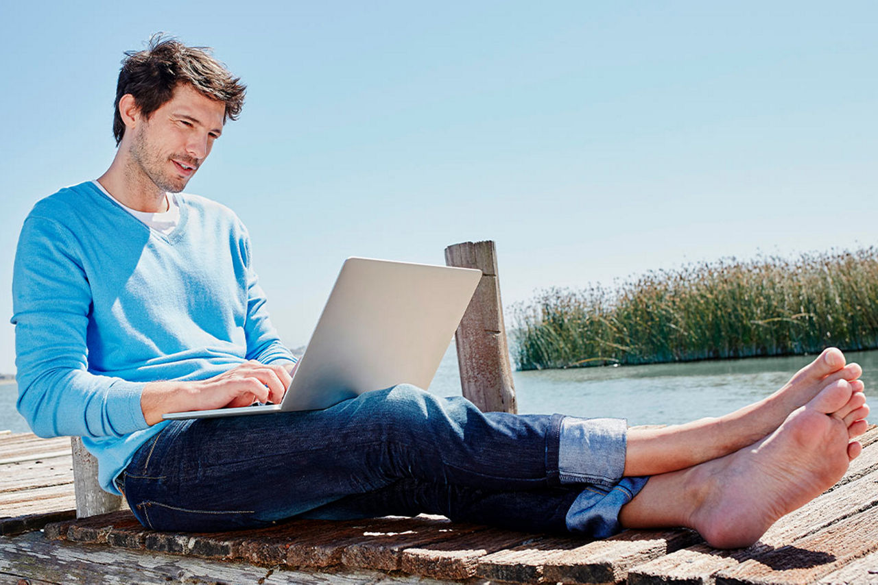 Ein Mann sitzt auf einem Steg am Wasser und hält sein Notebook in der Hand