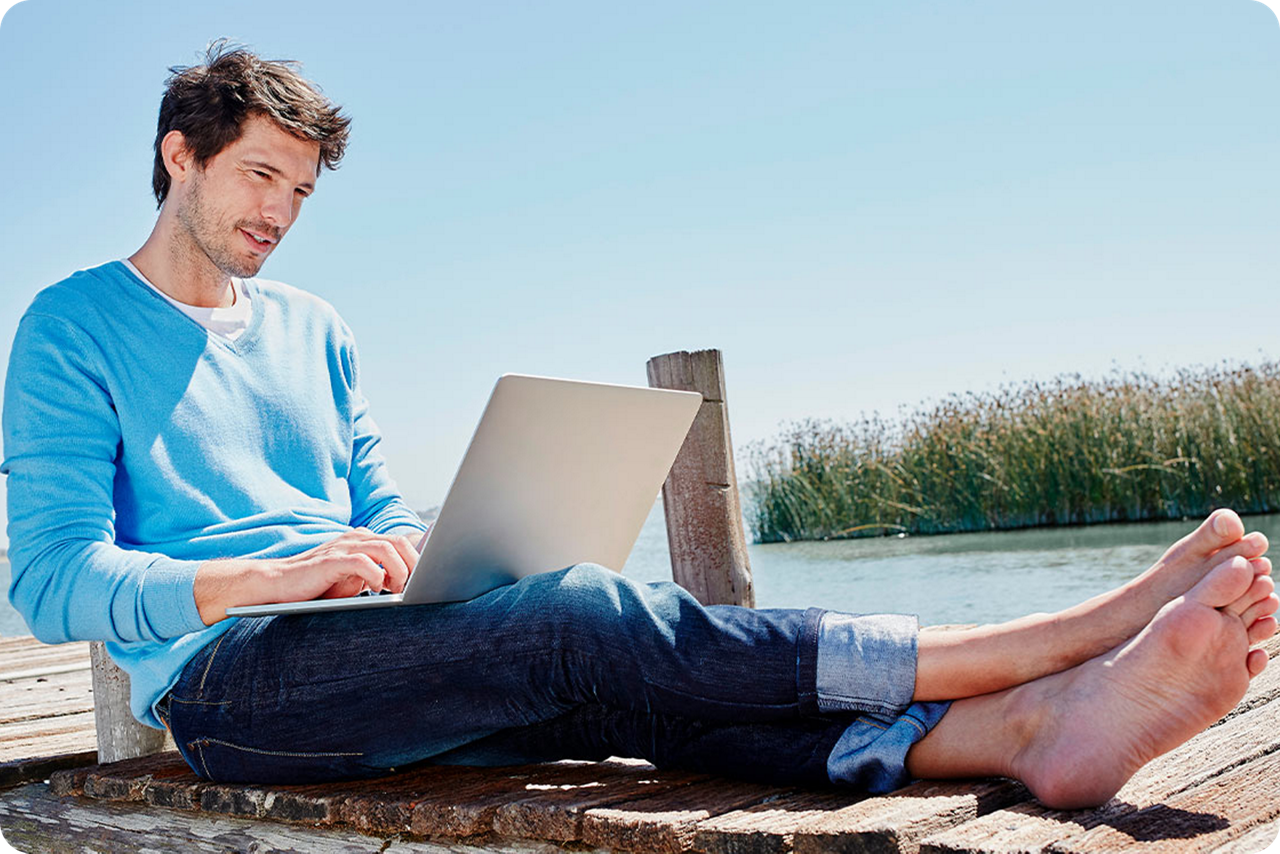 Ein Mann sitzt auf einem Steg am Wasser und hält sein Notebook in der Hand