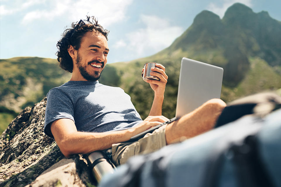 Mann sitzt in einer Gebirgslandschaft gemütlich mit einer Tasse am Laptop