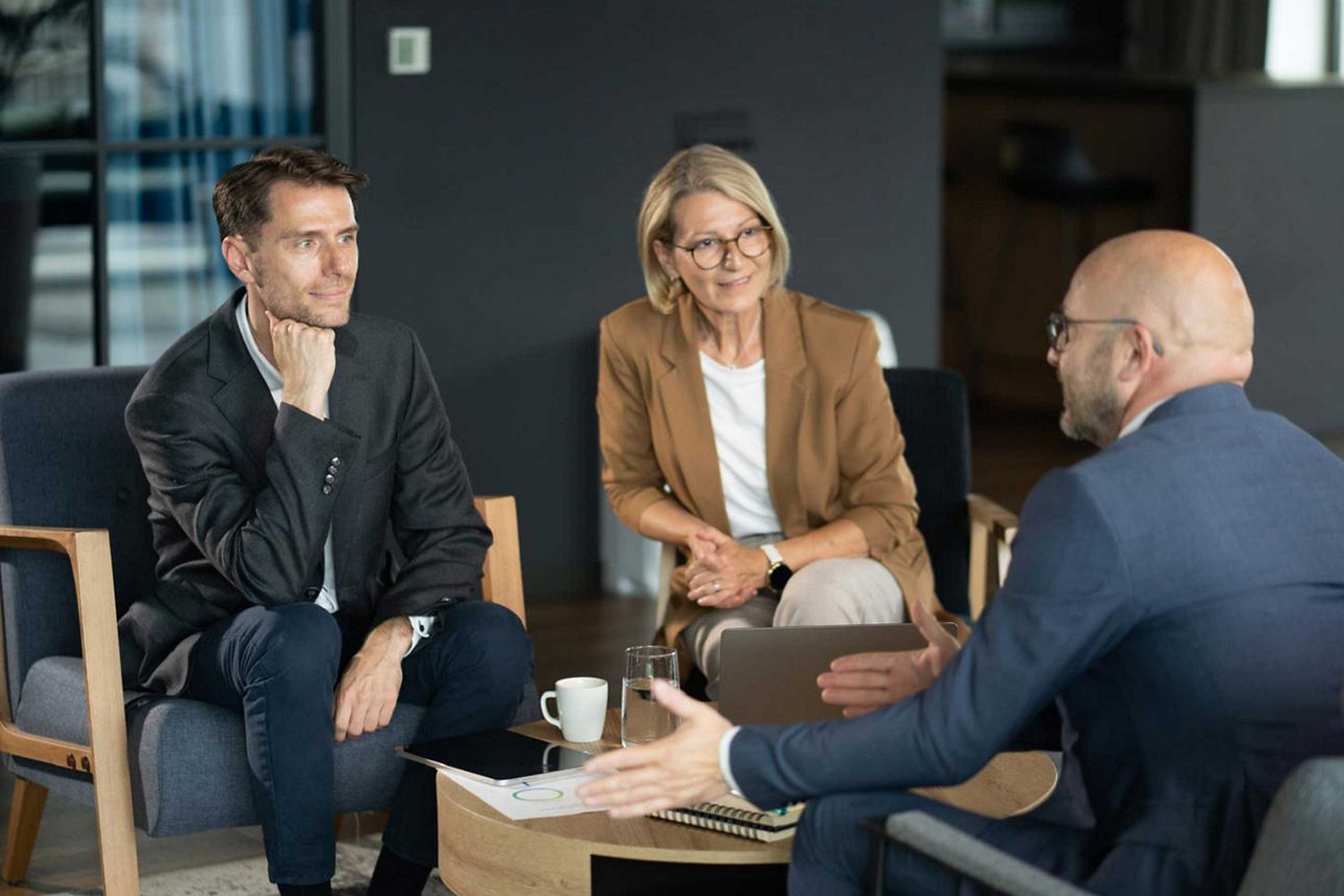  Drei Personen in einem engagierten Beratungsgespräch. Ein Berater erläutert einem Paar seine Anlagestrategie mithilfe von Un